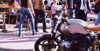 Moto aparcada en primer plano, y varias personas paseando o sentadas en una terraza en el fondo