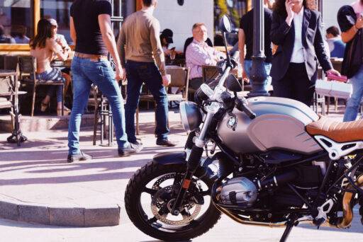 Moto aparcada en primer plano, y varias personas paseando o sentadas en una terraza en el fondo